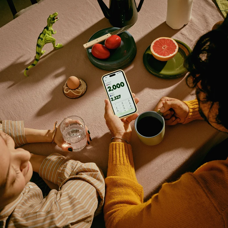 Two people at a breakfast table, with one holding a smartphone showing a currency conversion.