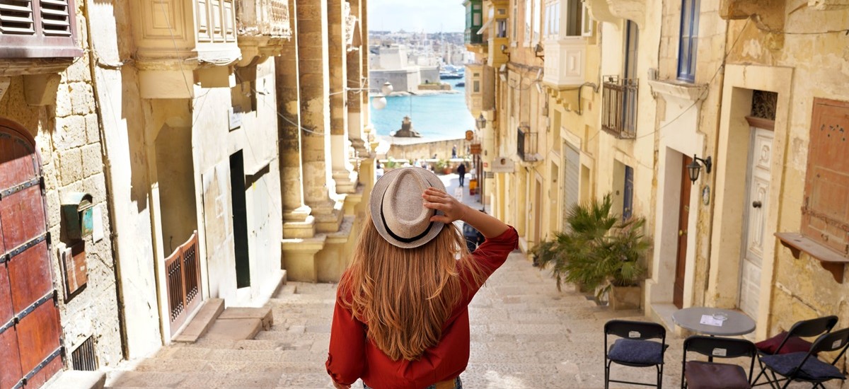 Tourist in orange shirt and hat walking away from camera through Maltese streets towards the harbour