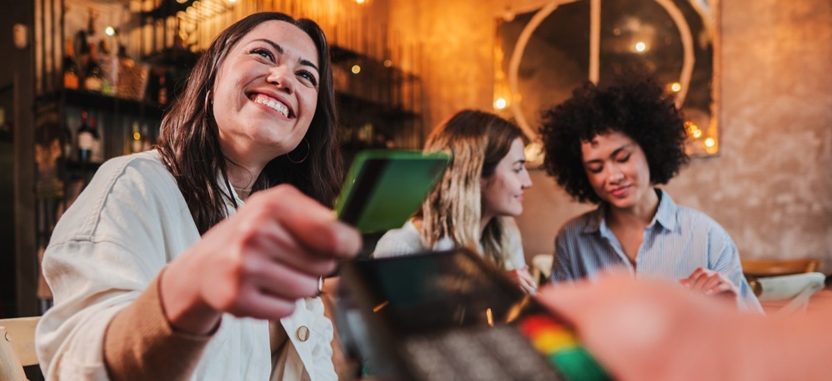 Smiling woman in a restaurant paying for a meal with a green card on a contactless reader