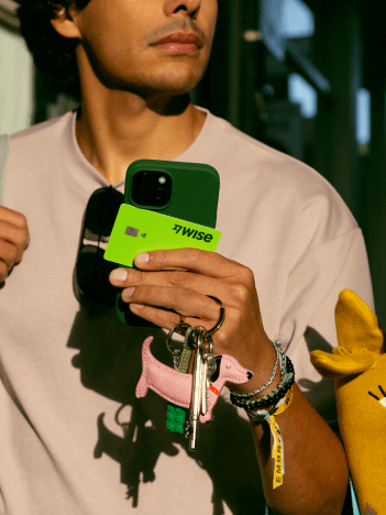 Un hombre camina por una calle comercial. Lleva su teléfono, gafas de sol y una tarjeta Wise en la mano.