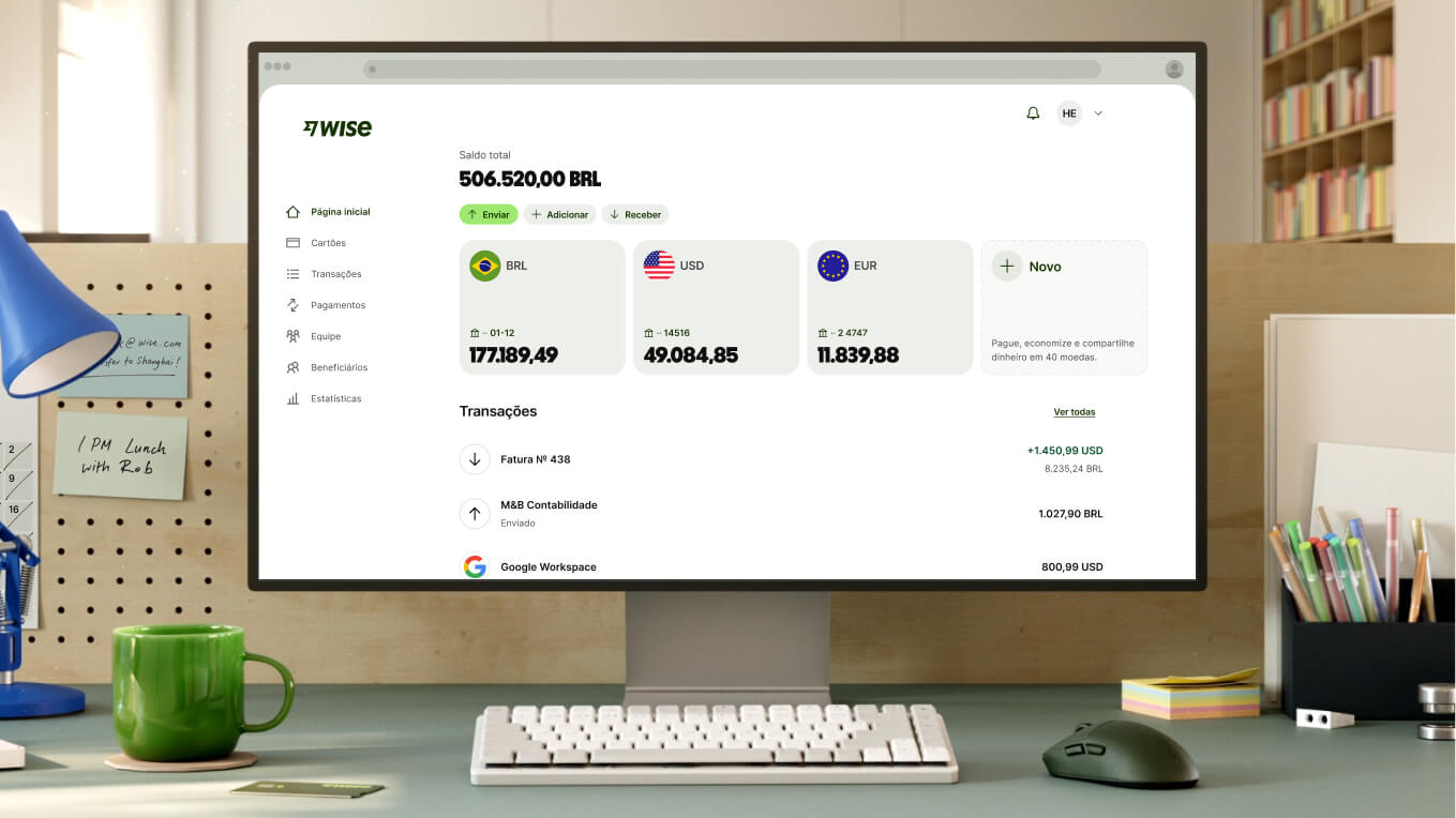 A imagem mostra de perto uma mesa de trabalho moderna, com um monitor branco exibindo um painel da Wise. A tela mostra um saldo total de "506.520,00 BRL" e saldos em três moedas diferentes: BRL com "177.189,49," USD com "49.084,85," e EUR com "11.839,88", seguido de um botão com a opção de abrir uma conta. As transações recentes são listadas abaixo. Na frente do monitor há um teclado compacto branco, um mouse cinza escuro e uma caneca de cerâmica em um tom de verde vivo e brilhante.